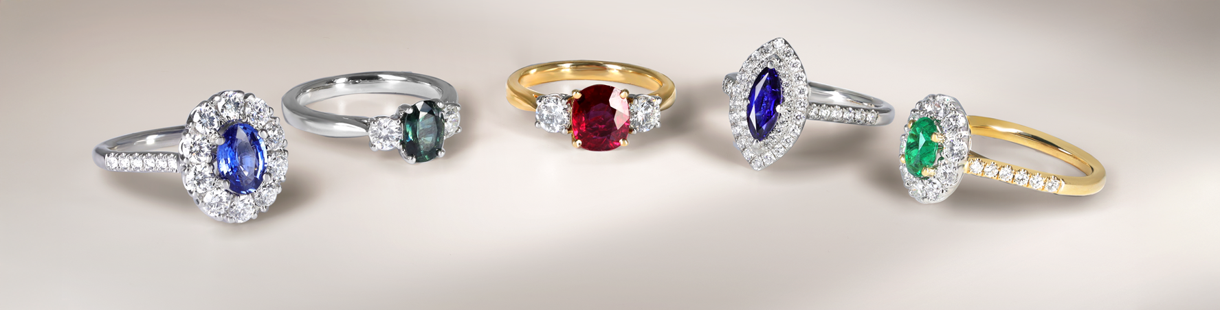 Neutral background with (left to right) oval sapphire cluster platinum ring, teal sapphire and diamond three stone platinum ring, oval ruby and diamond yellow gold three stone ring, marquise sapphire platinum band cluster, oval emerald cluster yellow gold ring